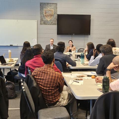 Students sit at tables, listening to a panel presentation.